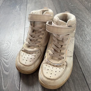 Beige höga sneakers med kardborreband - Säljer ett par beige höga sneakers med snörning och kardborreband upptill. Skorna har perforerade detaljer på tån och sidan samt platt sula. Perfekta för dig som gillar en sportig och avslappnad stil.