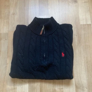 Svart stickad tröja med dragkedja från Polo Ralph Lauren - Kabelstickad svart tröja från Polo Ralph Lauren med halv dragkedja och röd broderad logga på bröstet. Tröjan har hög krage och långa ärmar, perfekt för kyligare dagar.