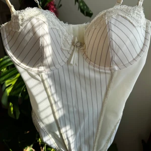 Vit korsett-topp med spets och rosett - Säljer ny en vit korsett-topp med tunna axelband, spetsdetaljer vid bysten och en liten rosett framtill från Ellos. Storlek 75C. Toppen har tunna vertikala ränder och en figurnära passform. Perfekt för att skapa en trendig och feminin look.