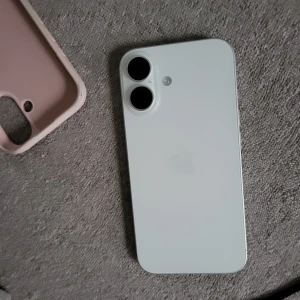 Vit iPhone 16  med rosa skal till Som gåva, jag tar 13900 kr för allt  - Säljer en vit iPhone med två stora kameralinser på baksidan. Telefonen har en stilren design och kommer med ett ljusrosa silikonskal. Skalet är mjukt och skyddar mobilen bra. Perfekt för dig som gillar enkel och modern stil.