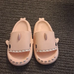 Rosa hajtofflor från Crayon friends - Säljer ett par gulliga rosa tofflor från Crayon Friends med hajdesign. Tofflorna har öppna tår, hajfenor på sidorna och tänder framtill. Perfekta för att liva upp vardagen med lite humor och färg. Inte användas många gånger.