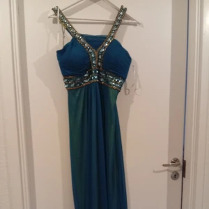 Blå långklänning med paljettdetaljer - Säljer en blå långklänning med breda axelband och glittriga paljettdetaljer längs byst och midja. Klänningen har en v-ringad framsida och är tillverkad i ett lätt, flytande material. Perfekt för bal eller fest.