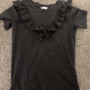 Svart t-shirt med volangdetalj - Svart t-shirt med korta ärmar och volangdetalj längs halsringningen. Enkel och stilren modell som passar till många olika outfits.💞