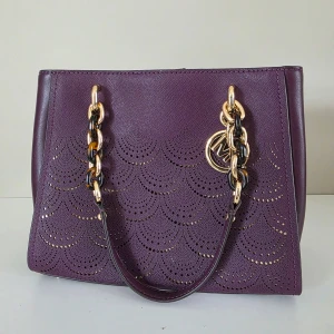 Lila handväska från Michael Kors med gulddetaljer - Lila handväska från Michael Kors med guldfärgade kedjehandtag och dekorativt MK-hänge. Väskan har ett perforerat mönster framtill och flera fack inuti, inklusive ett med dragkedja. Se profil för fler väskor i nytt/mycket bra skick