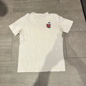 Vit t-shirt med färgglatt tryck från Nike - Vit t-shirt från Nike med ett litet färgglatt tryck på bröstet och ett större tryck på ryggen. Klassisk passform och rund halsringning. Perfekt för en avslappnad streetwear-look. M men passar även S.