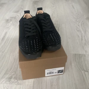 Loubs - Säljer ett par svarta sneakers med coola svarta nitar på tån och ovandelen. Skorna har snörning och en unik röd sula som sticker ut. Perfekta för dig som vill ha något extra i din stil!