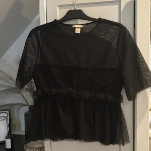 Svart transparent meshblus med volang - Säljer en svart blus i transparent mesh med korta ärmar och volangdetaljer vid midjan och bröstet. Perfekt för att skapa en trendig lager-på-lager-look. Blusen har rund halsringning och luftig passform.