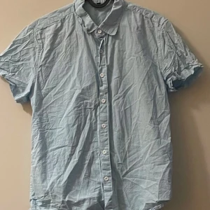 Ljusblå kortärmad skjorta - En ljusblå skjorta med korta ärmar och klassisk krage. Skjortan har knäppning framtill med vita knappar och är tillverkad i ett lätt material, perfekt för varmare dagar.