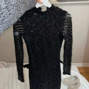 Svart glittrig långärmad klänning - Säljer en svart långärmad klänning med glittriga paljetter och transparenta partier på ärmar och axlar. Klänningen har en tight passform och hög hals, samt en snygg öppen rygg. Perfekt för dig som vill sticka ut på festen!