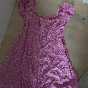 Rosa blommig klänning med knappar - Söt rosa klänning med små vita blommor och knappar framtill. Klänningen har korta puffärmar och en lätt rynkad urringning. Perfekt för sommarens alla tillfällen!