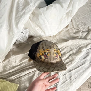 Gucci keps med tigertryck - Säljer en beige och svart keps från Gucci med klassiskt GG-mönster och två tigrar framtill. Kepsen har svart mesh på baksidan och justerbar passform. Perfekt för dig som vill sticka ut med en lyxig accessoar.
