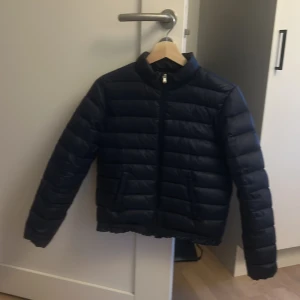 Mörkblå dunjacka från Polo Ralph Lauren - Säljer en mörkblå dunjacka från Polo Ralph Lauren med klassisk broderad logga på bröstet. Jackan har dragkedja framtill, ståkrage och två fickor. Perfekt för kyligare dagar och enkel att matcha med olika outfits.