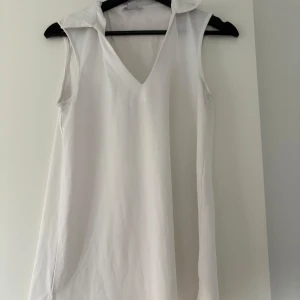 Vit ärmlös topp med krage  - Säljer en stilren vit ärmlös topp från Amisu i storlek XS. Toppen har en djup v-ringning och en klassisk krage, vilket ger en snygg och modern look. Perfekt att matcha med jeans eller kjol!