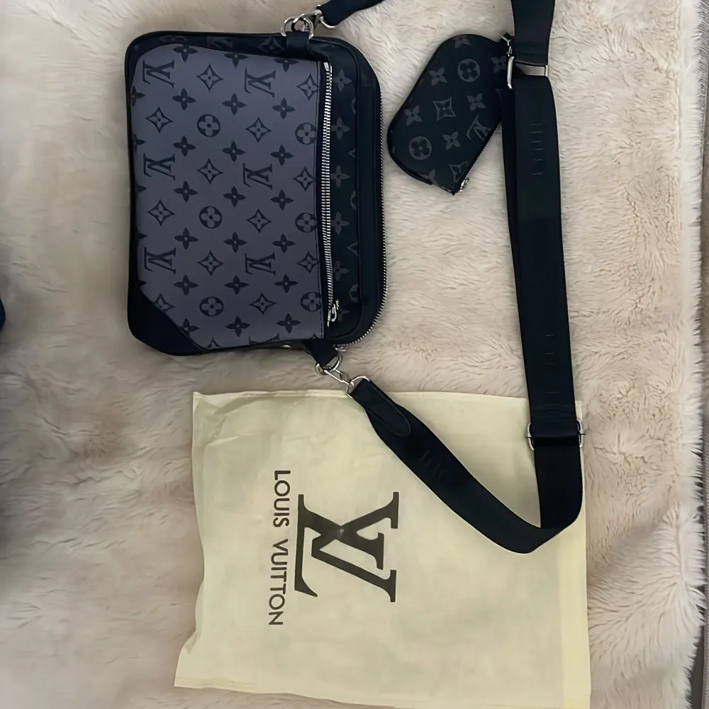 Säljer en svart axelväska med klassiskt monogrammönster i grått. Väskan har justerbar axelrem, silverfärgad dragkedja och medföljande liten rund accessoar. Perfekt för att bära dina viktigaste saker med stil.. Laukut & Käsilaukut.