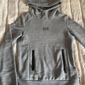 Grå hoodie från EA7 Emporio Armani - Säljer en stilren grå hoodie från EA7 Emporio Armani med svarta dragkedjor på fickorna och logga på bröstet. Tröjan har lång ärm och huva, perfekt för en sportig look.