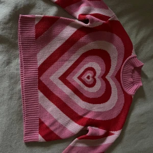 Rosa och röd stickad tröja med hjärtmönster - Superfin stickad tröja i olika nyanser av rosa och rött med stort hjärtmönster på framsidan. Tröjan har rund halsringning och långa ärmar. Aldrig använd!