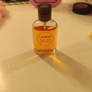 La Rive Touch Woman Eau de Parfum - En elegant parfymflaska med La Rive Touch Woman Eau de Parfum. Flaskan är genomskinlig med rundade kanter och innehåller en gyllene vätska. Svart lock och enkel, modern design med lila text på etiketten.