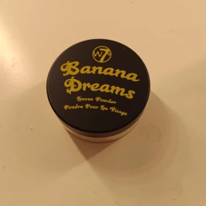 Banana Dreams Loose Powder från W7 - Banana Dreams är ett löspuder från W7 i en rund burk med svart lock och gul text. Pudret är ljusgult och används för att fixera smink och ge en matt finish. Perfekt för att ljusa upp under ögonen eller hela ansiktet. Endast använd en gång. ❤️