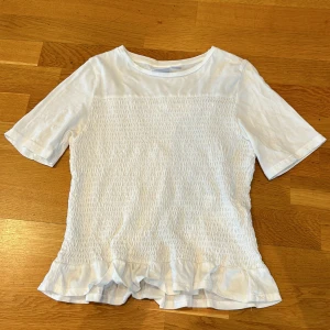 Vit topp med volang från Gina Tricot - Säljer en söt vit topp från Gina Tricot med korta ärmar och smockad framsida och baksida. Toppen har volangdetalj längst ner och är tillverkad i mjuk bomull. Perfekt för en fräsch och stilren look!