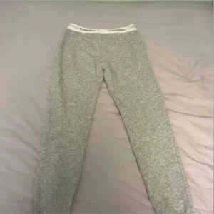 Grå mjukisbyxor från Calvin Klein - Grå mjukisbyxor från Calvin Klein med vit resår i midjan där loggan syns tydligt. Byxorna har en rak passform och är tillverkade i mjukt material, perfekta för en avslappnad stil. Har även ett par svarta till salu 