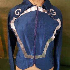 Blå jacka med silvriga detaljer - Snygg blå jacka med silvriga vågformade detaljer och dragkedja framtill. Jackan har krage och lång ärm, samt dekorativa G-bokstäver på axlarna. Perfekt för dig som vill sticka ut med en unik stil.