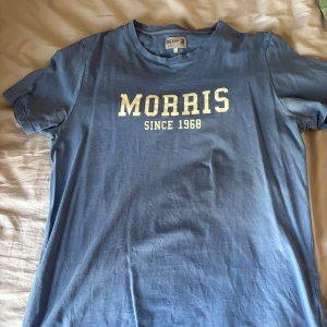 Blå t-shirt från Morris - Säljer en blå t-shirt från Morris med trycket 'MORRIS SINCE 1968' i vitt på bröstet. Klassisk rund halsringning och korta ärmar. Perfekt för en avslappnad stil.
