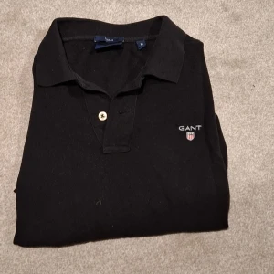 Svart pikétröja från GANT - Klassisk svart pikétröja från GANT med broderad logga på bröstet. Tröjan har krage, knappar framtill och är tillverkad i mjuk bomull. Perfekt för en stilren och avslappnad look. Säljes pga något kort för mig i stl M 