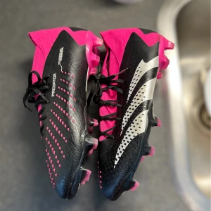 Adidas Predator fotbollsskor i svart och rosa - Säljer ett par Adidas Predator fotbollsskor i svart och rosa med vita detaljer. Skorna har snörning och mönstrad ovansida för extra grepp på planen. Perfekta för dig som vill sticka ut på fotbollsplanen!