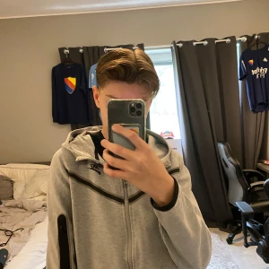 Grå hoodie från Nike med dragkedja - Säljer en grå hoodie från Nike med dragkedja framtill och svart logga på bröstet. Tröjan har huva, långa ärmar och en praktisk ficka med dragkedja på ena ärmen. Perfekt för en sportig och avslappnad stil.