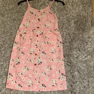 Rosa blommig klänning med knappar - Söt rosa klänning med vita och gula blommor, smala axelband och knappar framtill. Klänningen har en avslappnad passform och är perfekt för varma dagar. Fickor framtill och lätt, mjukt material.