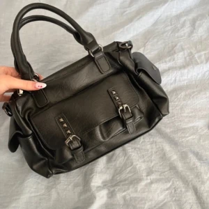 Svart handväska med nitar - Snygg Sthlm stil svart handväska i skinn med coola nitar och spännen på framsidan. Väskan har både handtag och en justerbar axelrem för olika bärstilar. Perfekt för att ge din outfit en edgy touch. Fickor på bägge sidorna!❤️
