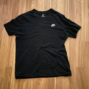 Svart t-shirt från Nike - Svart t-shirt från Nike med klassisk logga på bröstet. Enkel och stilren design med rund hals och korta ärmar. Perfekt för en sportig och avslappnad look.