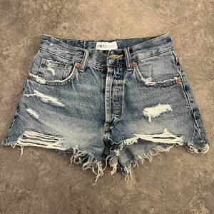 Blå slitna jeansshorts från ZARA - Riktigt snygga blå jeansshorts från ZARA, midrise och råa, fransiga kanter. Shortsen har slitningar och hål för en cool, avslappnad look. Klassisk femficksmodell med knappar i gylfen.