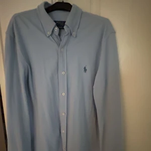 Ljusblå skjorta från Polo Ralph Lauren - Säljer en klassisk ljusblå skjorta från Polo Ralph Lauren med button-down krage och broderad logga på bröstet. Skjortan har långa ärmar och knäppning framtill. Perfekt till både jeans och chinos.