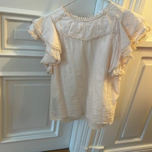 Vit blus med volangärmar och spetsdetaljer - Säljer en söt vit blus med korta volangärmar och fina spetsdetaljer längs kanterna. Blusen har en lös passform och rund halsringning, perfekt för en somrig look.