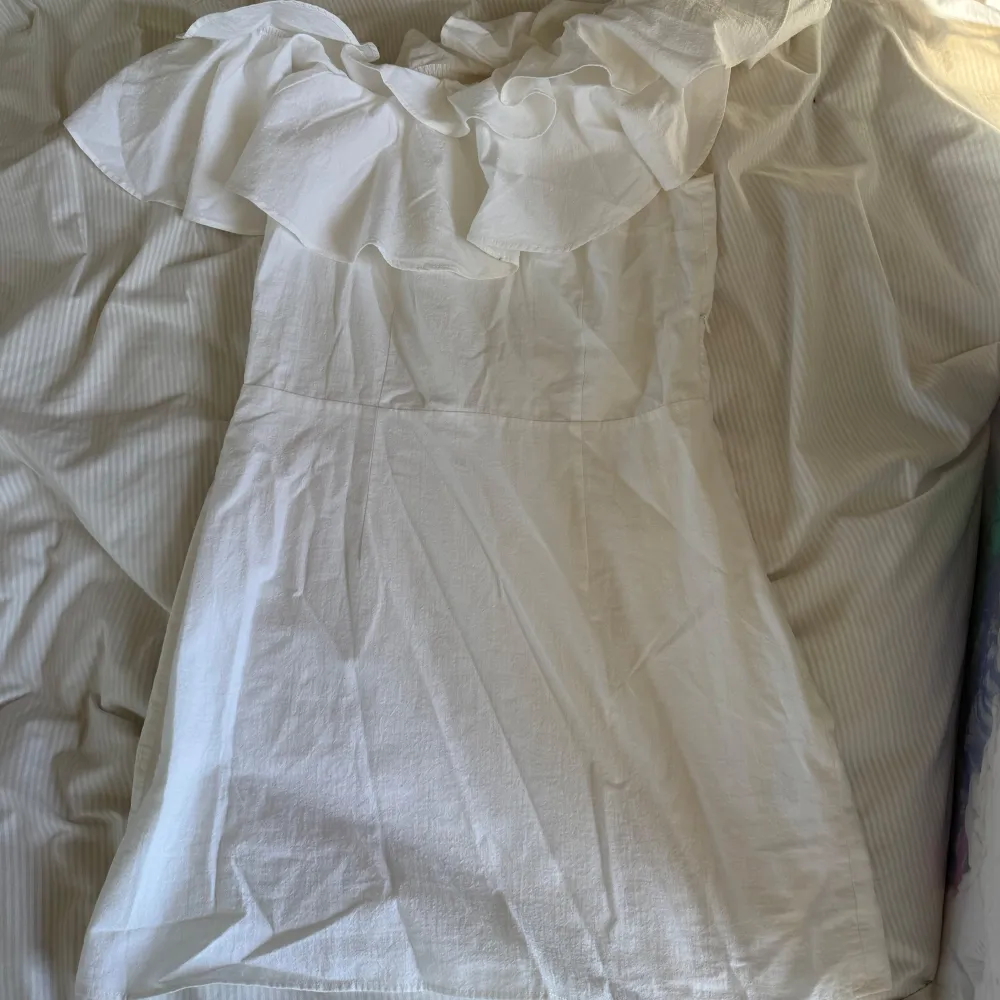 Säljer en vit kortklänning med offshoulder och stor volangdetalj över axlarna. Klänningen har en figurnära passform och dragkedja i sidan som har gått sönder i sömmarna, men väldigt lätt att sy. Perfekt för studenten eller sommarens tillfällen när du vill sticka ut lite extra.. Mekot.