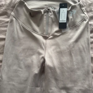 Beige träningstights  - Säljer ett par beige träningstights från Aim'n med hög midja och diskret logga på höften. Tightsen har en tight passform och är tillverkade i ett stretchigt syntetmaterial som passar perfekt för gym eller yoga.