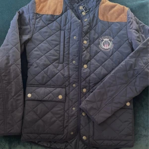 Quiltad jacka från Lexington med mockadetaljer - Snygg quiltad jacka från Lexington i mörkblått med bruna mockadetaljer på axlarna. Jackan har guldiga knappar, dragkedja, ståkrage med spänne och två stora fickor framtill. Broderad Lexington-logga på bröstet.