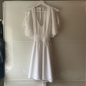Vit klänning med volangärmar och v-ringning - En fin vit klänning med v-ringad hals och volangiga, korta ärmar. Perfekt till sommaren, studenten eller konfirmationen. Från Bubbleroom. Mycket bra skick. 