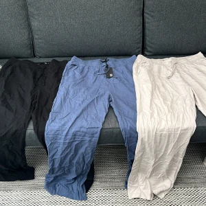 Tre par linnebyxor i svart, blått och beige - Tre par luftiga linnebyxor i svart, blått och beige. Byxorna har resår i midjan och raka ben, perfekta för varma dagar. Snygga att matcha med en enkel topp eller skjorta.