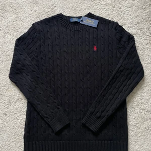 Kabelstickad tröja från Ralph Lauren - Säljer en svart kabelstickad tröja från Ralph Lauren med rund halsringning och röd broderad logga på bröstet. Tröjan har långa ärmar och ribbade muddar vid ärmslut och nederkant. Perfekt för en stilren look. (Lapp finns men sitter ej på. Tröjan är bara använd någon enstaka gång).