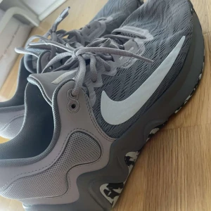 Grå Nike sneakers med mönstrad sula - Säljer ett par grå Nike sneakers med vit swoosh och coolt svartvitt mönster på sulan. Skorna har snörning och är tillverkade i syntetmaterial med mesh för extra andningsförmåga. Perfekta för dig som gillar sportig stil.
