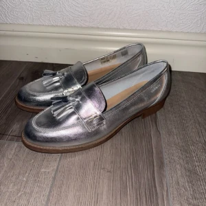 Silvriga loafers med tofsar - Säljer ett par snygga silvriga loafers från Unisa med dekorativa tofsar framtill. Skorna har en låg klack och rund tå, samt en platt sula i beige. Perfekta för dig som vill sticka ut med metallic-look!