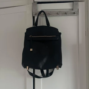 Svart ryggsäck - Snygg svart ryggsäck i skinnimitation med guldfärgade detaljer och dragkedja framtill. Väskan har både handtag och justerbara axelremmar samt flera fack inuti för enkel organisering. Perfekt för dig som vill ha en stilren och praktisk accessoar.
