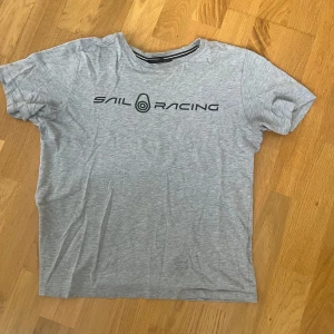Sail Racing T-shirt  - Snygg grå t-shirt från Sail Racing med svart logga tryckt på bröstet och en mindre logga i nacken. Klassisk rund halsringning och korta ärmar. Skön och lätt i materialet, perfekt för en avslappnad stil.