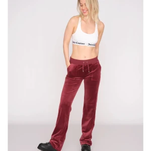 Vinröda velourbyxor Juicy Couture - Säljer ett par vinröda velourbyxor från Juicy Couture med bred resår i midjan och snörning. Byxorna har raka ben, stora fickor fram och bakfickor med lock och silverfärgade knappar. Perfekta för en chill och trendig look.