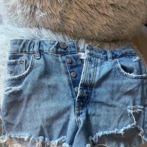 Blå jeansshorts med fransar från Zara - Snygga blå jeansshorts från Zara med hög midja, slitna detaljer och fransig kant nedtill. Klassisk femficksmodell med knappar framtill och coolt avslappnat vibe. Perfekta för varma dagar och en chill stil.