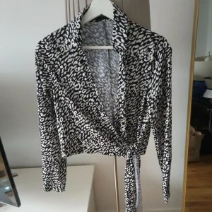 Leopardmönstrad omlottopp svart/vit - Snygg omlottopp med svart och vitt leopardmönster. Toppen har långa ärmar, krage och knytband i midjan för en cool och trendig look. 