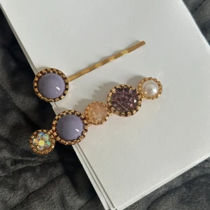 Guldiga hårspännen med pärlor & stenar - Två hårspännen i guldton med runda dekorationer i lila, pärlvit och glittrande stenar. Ena spännet har fem olika stenar, bland annat lila, pärlemor och glittrig, medan det andra har en lila sten. Perfekt för att ge frisyren en färgglad och lyxig touch.