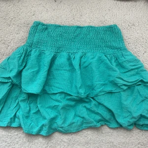 Turkos volangkjol med små prickar - Supersöt turkos kortkjol med volanger och små vita prickar. Kjolen har bred resår i midjan för skön passform och är perfekt för varma dagar. Lätt och luftig känsla, passar dig som gillar färg och lekfull stil.